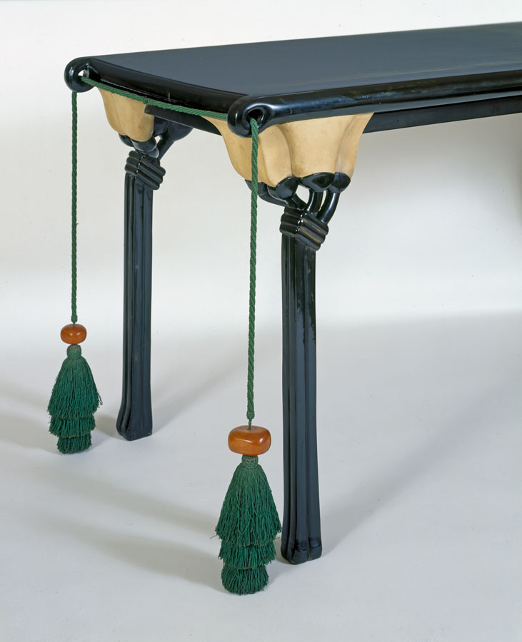 Detail view of a blue lacquered table with lotus flower legs and large green tassels hanging from the short edge.