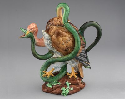 Majolica vulture and serpent shaped teapot, green serpent with pink, brown, black, white, and yellow culture standing on brown and green rock.