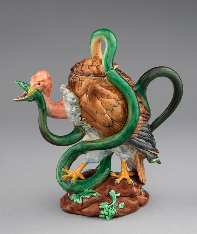 Majolica vulture and serpent shaped teapot, green serpent with pink, brown, black, white, and yellow culture standing on brown and green rock.