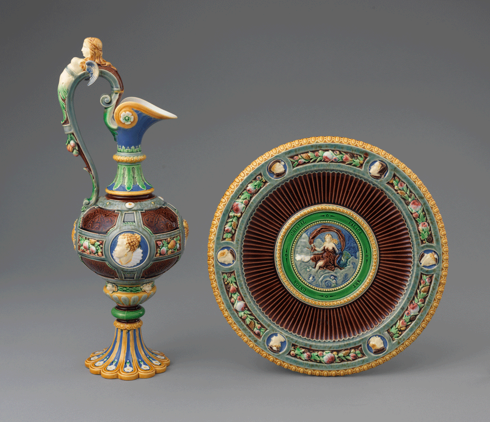 Majolica ewer and stand. Ewer features a cameo of Queen Elizabeth I and a mermaid handle in dark red, yellow, blue, and green. Stand features a robed woman in the center with a border of floral friezes and cameos around the edge in matching colors to the ewer.