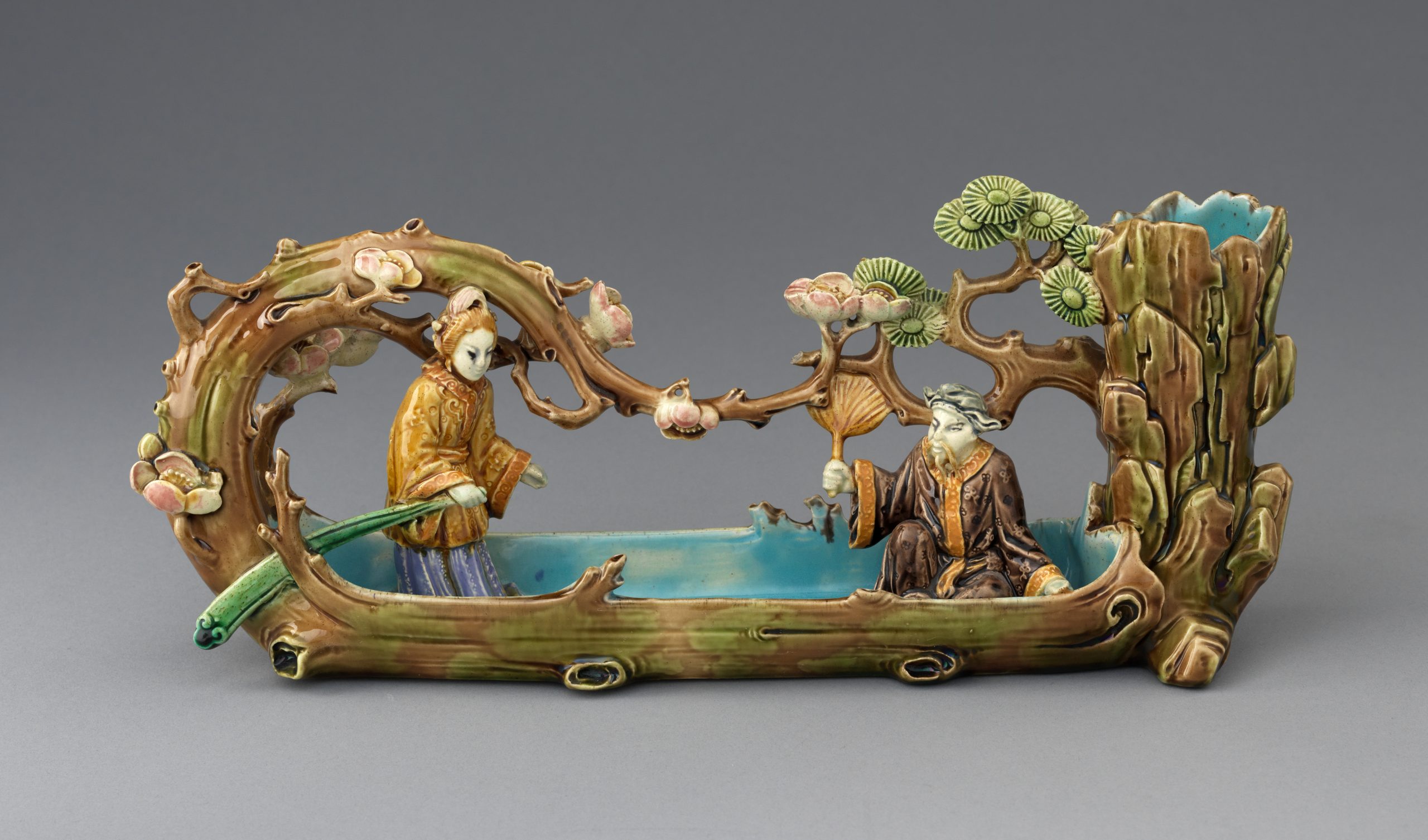 Small majolica sculpture of "Japanese" boatmen in long boat made to look like a carved out tree trunk with pink flowering branches curving up and over each side.