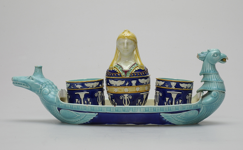 Egyptian revival majolica desk set and tray. Canopic jar style pot and two wells with dark blue ground, gold detailing, and white figures, lid in shape of Egyptian bust. Boat-shaped tray with darl blue ground and turquoise figures in shape of crocodile and bird.