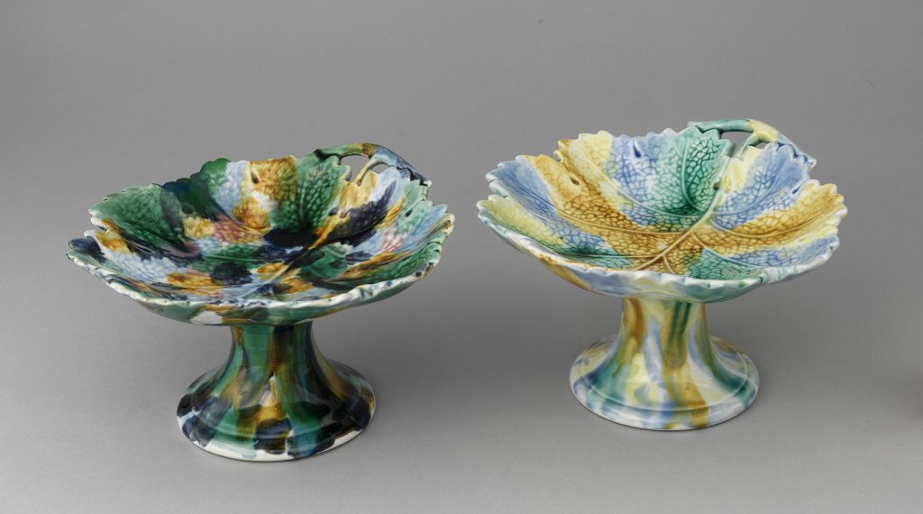 Two majolica leaf bowls on stands, body of various spots of green, blue, and yellow.