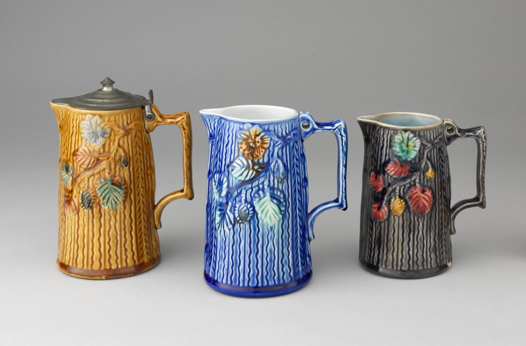 Three majolica jugs with wood-texture body and leafy branch motif. Left jud is brown, middle jug is blue, right jug is gray.