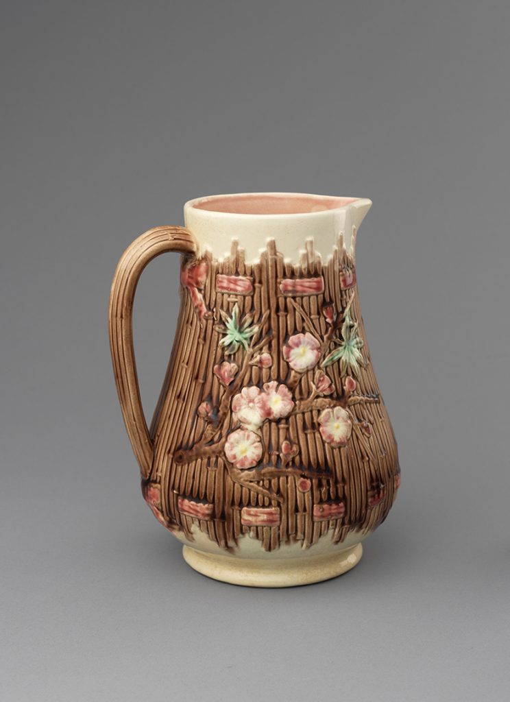 Majolica tea jug with reed texture on body and handle. Base and mouth have cream colored ground, body and handle have brown ground. Body has brown flower branch with pink flowers and green leaves.