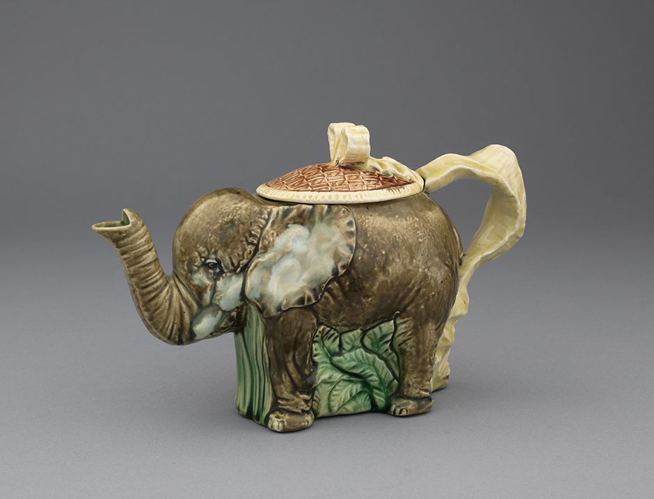 Majolica teapot in the form of an elephant with yellow ribbon handle. Brown ground on elephant with green foliage and turtle shell motif lid.
