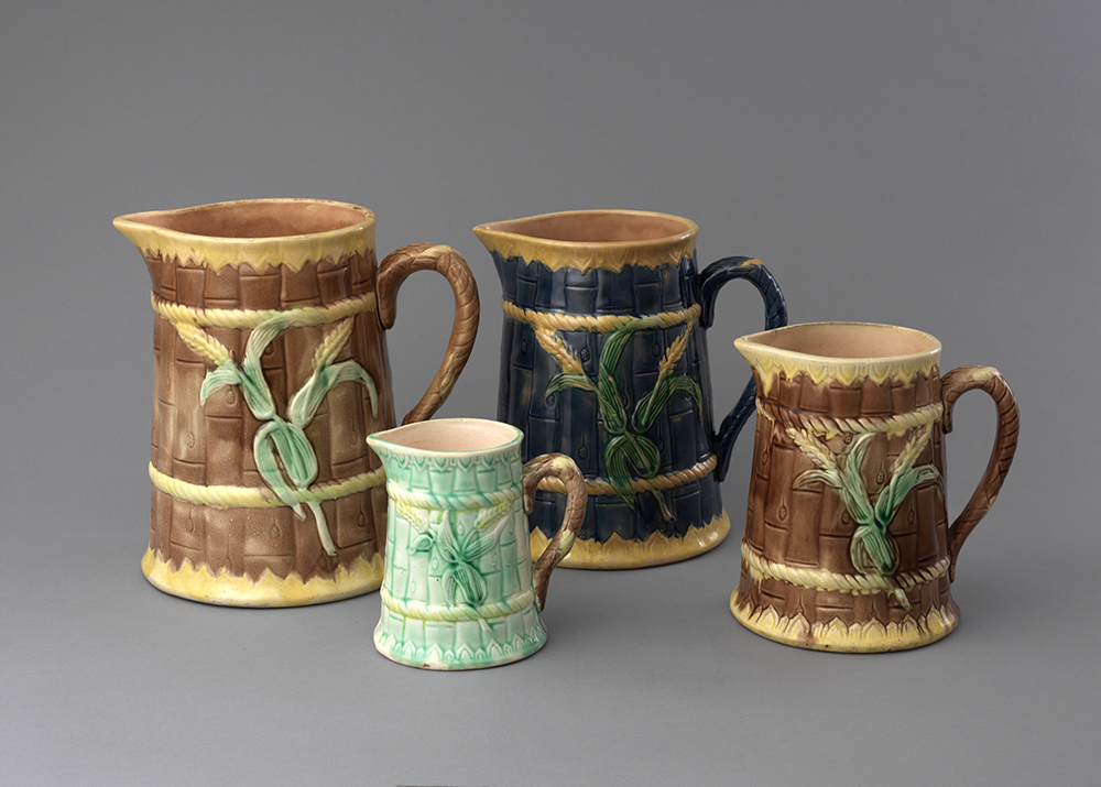 Four majolica jugs with bamboo texture ground and handles. Leftmost large jug and rightmost small jug have brown ground with yellow detailing. Small left jug was turquoise ground with brown handle and center jug has dark blue ground and handle with yellow detailing. All four jugs have yellow sheaf of wheat with green leaves on body.