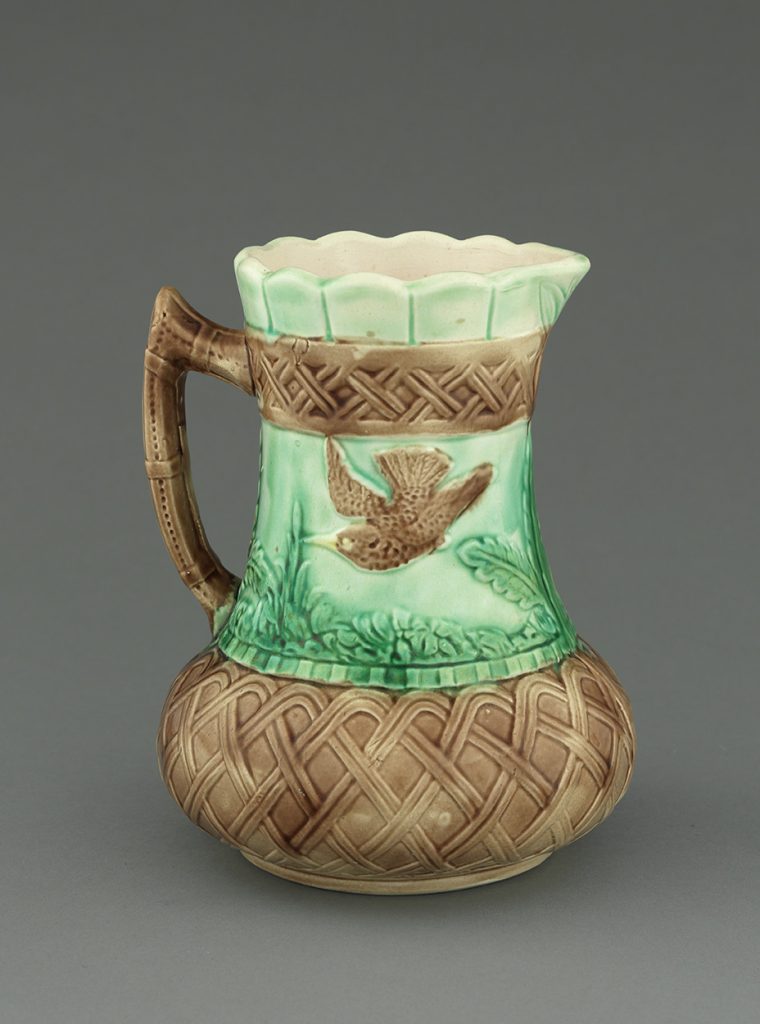 Majolica jug with brown latticework on neck, base, and handle. Brown bird and green landscape background on center.