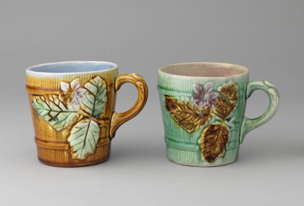 Two majolica mugs with wooden barrel body motif and leves and flower motif. left mug is brown with green leaves, right mug is green and brown leaves.