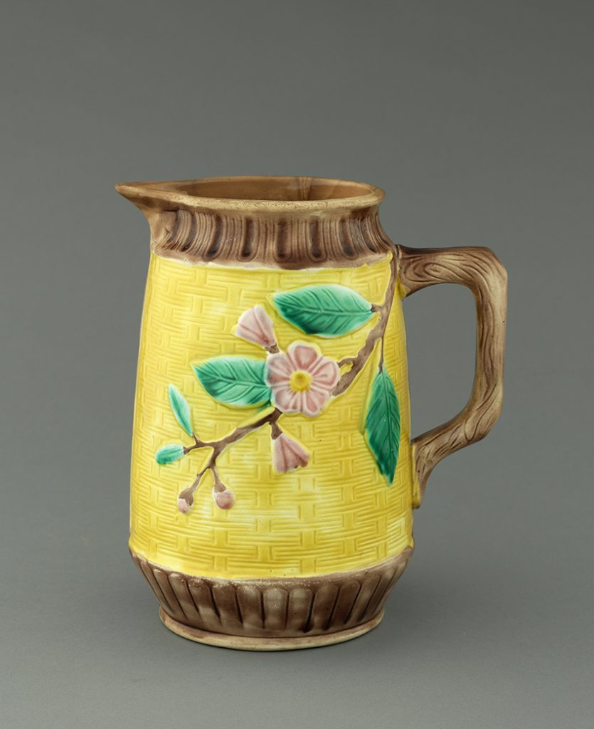 Majolica jug with yellow woven texture ground on body and brown wood texture ground on mouth, base, and handle. Body has brown branch with pink flower and buds and green leaves.