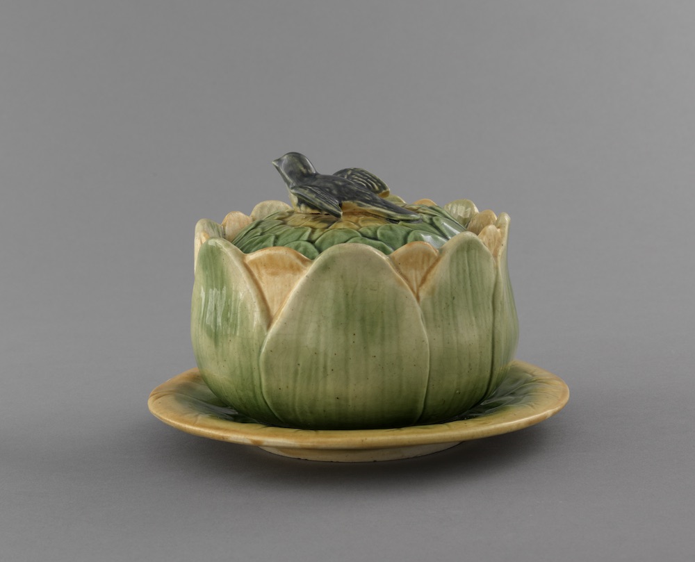 Green and yellow majolica butter dish and plate in the shape of an artichoke. Leaves overlap around artichoke center, dark green bird-shaped finial. finial l