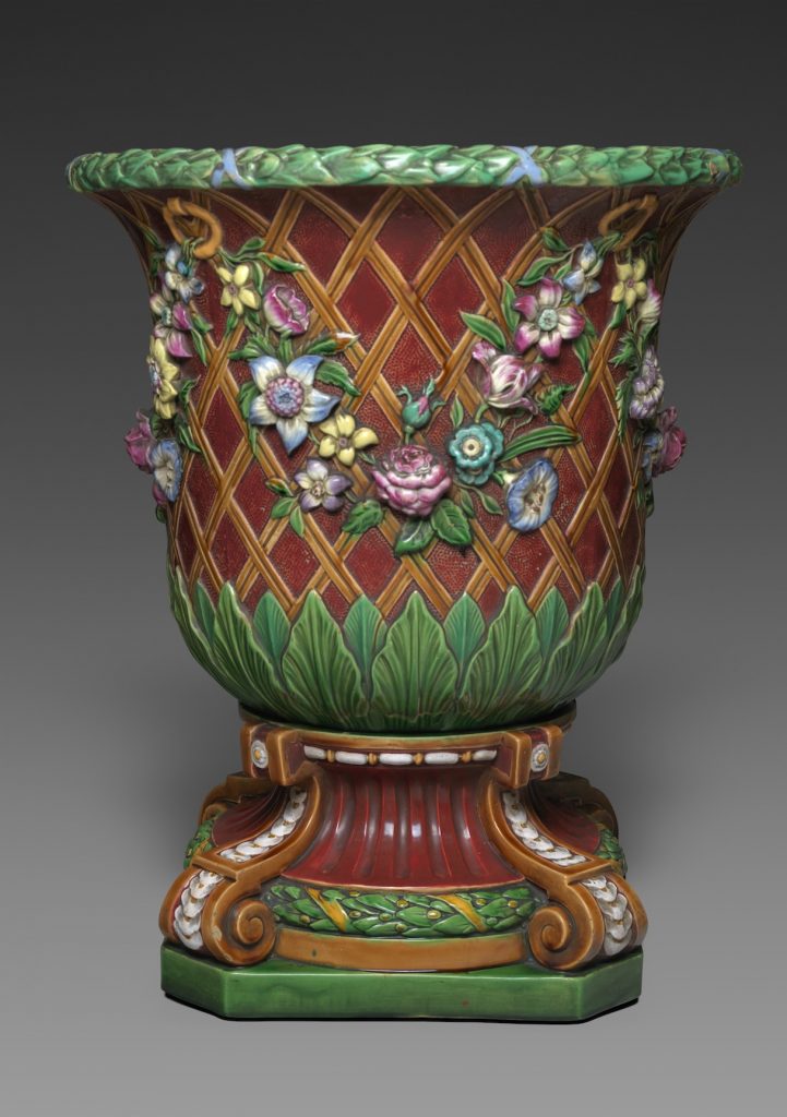 Majolica garden pot with flared mouth on volute pedestal. Body is brown latticework with festoon of pink and yellow flowers, base is green leaves and lip is green and yellow garland with pink ribbons.