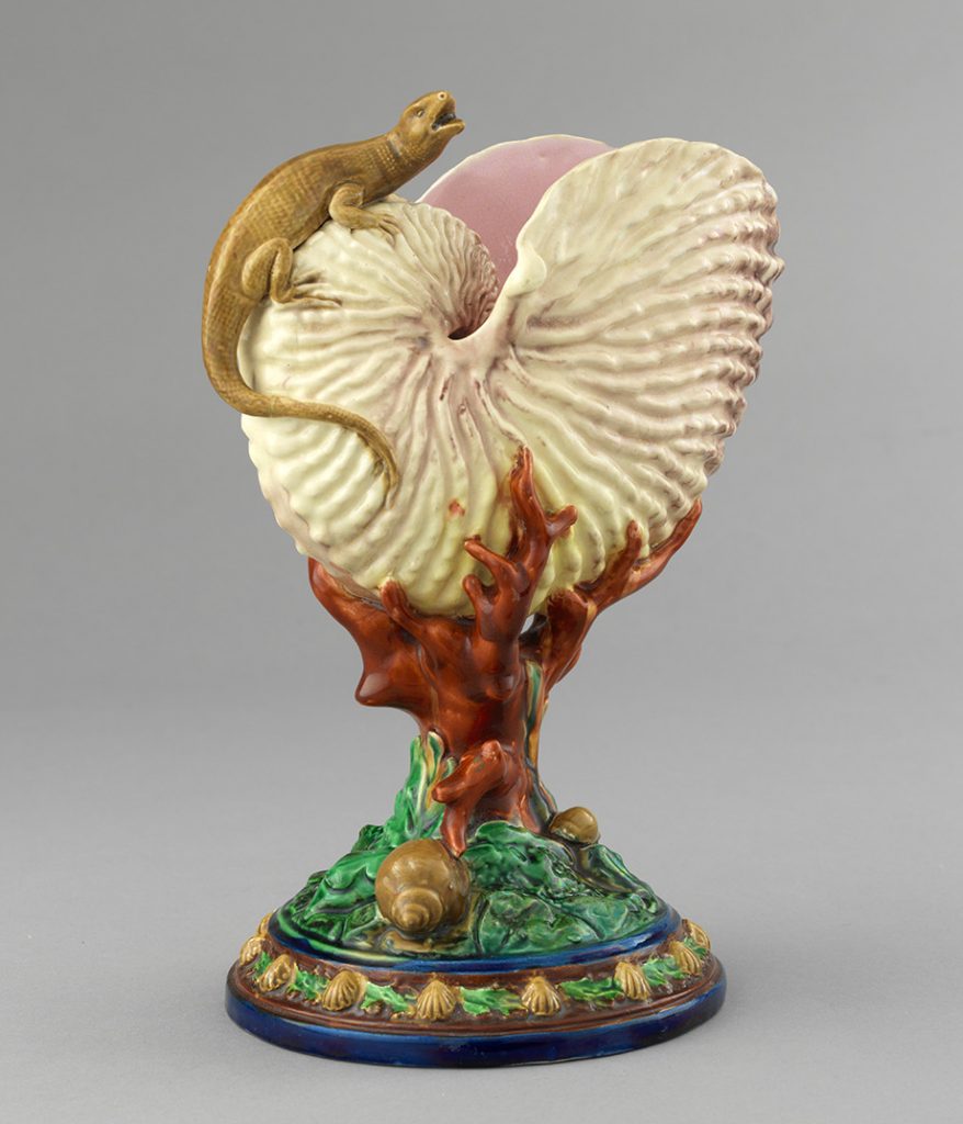 Majolica cup in the shape of a nautilus shell with lizard figure and coral stem with foliage and snail shells. White and pink shell with brown lizard, red coral, and brown and green base.