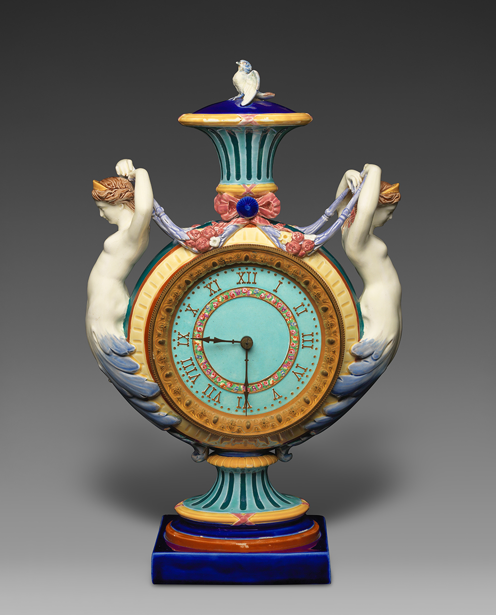 Majolica clock vase with two mermaid figures and bird finial. Color palette of turquoise, dark blue, yellow, white, brown, and red.