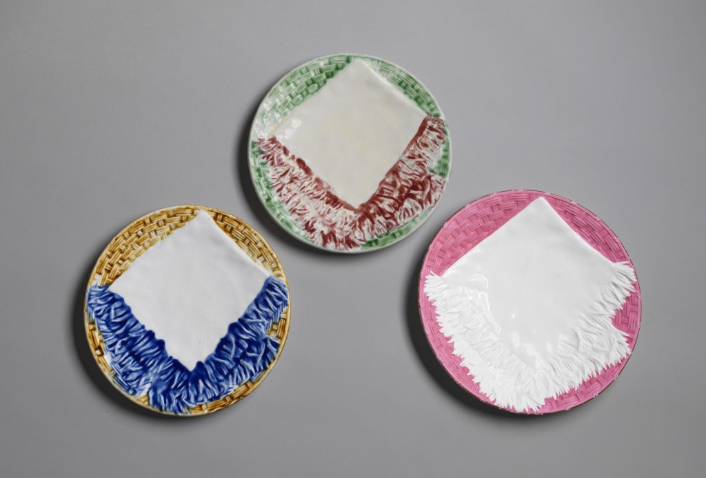 Three majolica plates with folded napkin motif, white napkins with colored or white fringe and grounds of various colors.