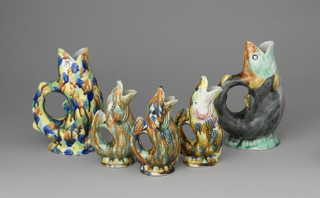 Set of five majolica fish jugs, three with fish sdale texture. Color palette of various spots of blue, yellow, green, and brown. Two end jugs are larger than three middle jugs.