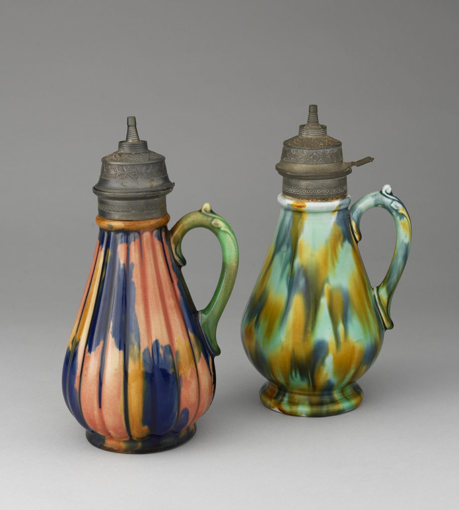 Two majolica ewers with fluting and vine handle and metal lids Color palette of various streaks of blue, green, red, and yellow.