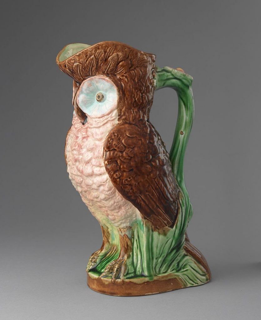 Majolica jug in the shape of an owl with pink body and brown head, wings, and feet. Green vine motif handle and base.