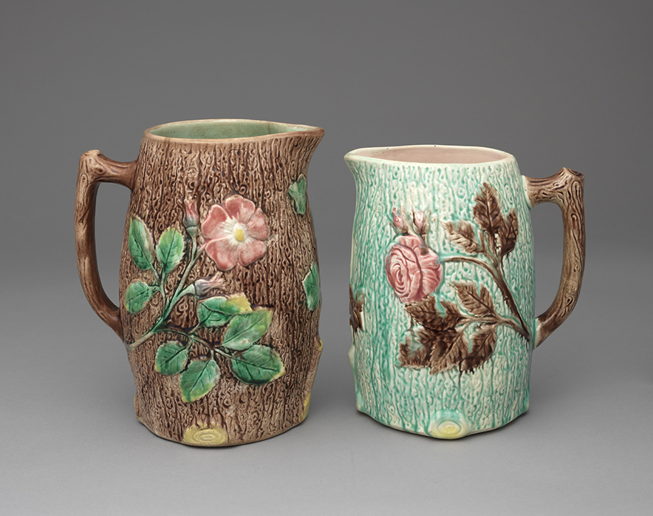 Two majolica jugs with wood-grain texture on body and handle. Left jug has brown ground with pink flowers and green leaves, right jusg has turquoise ground with pink flowers and brown handle and leaves.