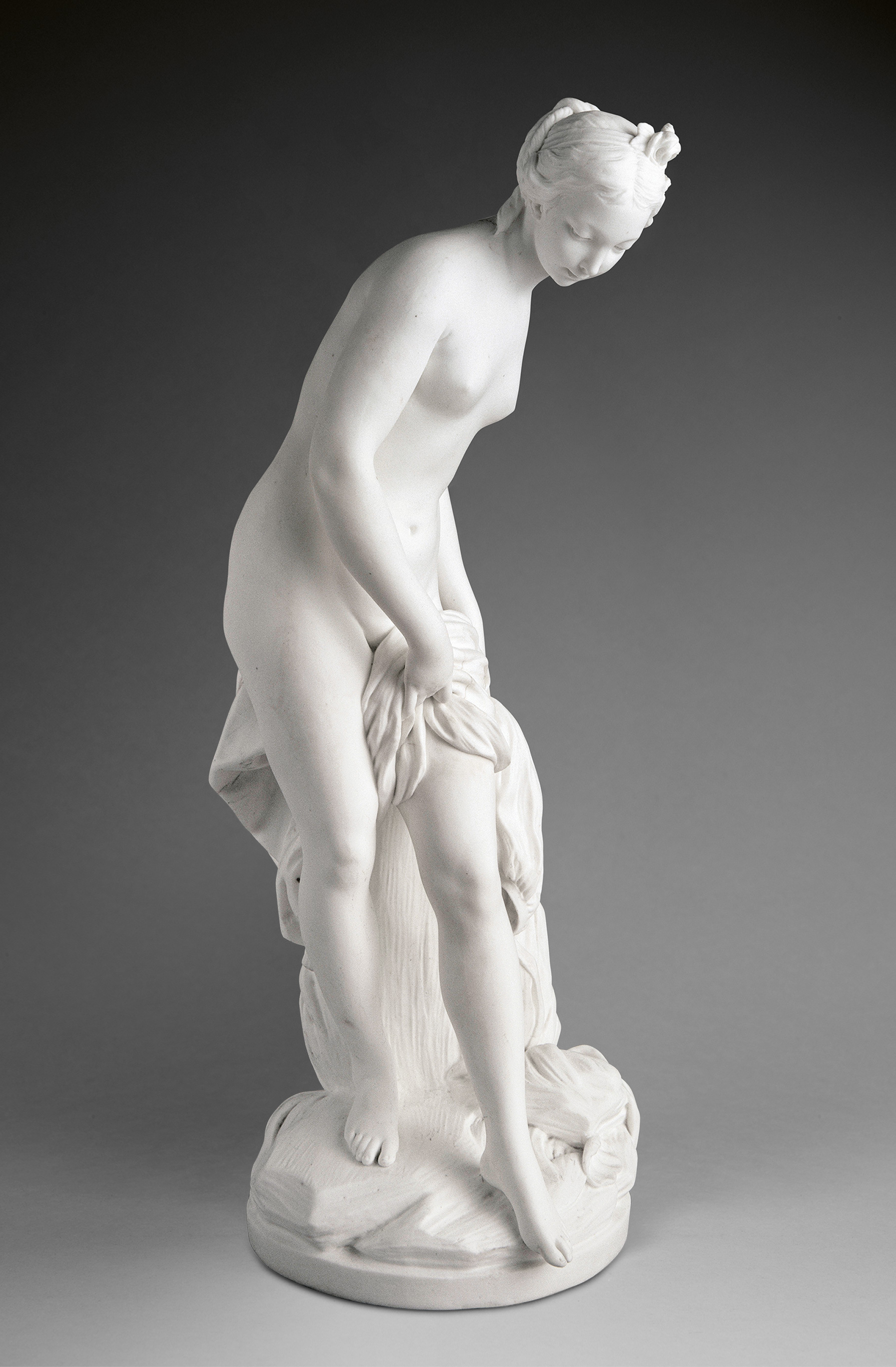 A white marble statue depicts a nude woman standing on a rounded base, gracefully bending forward while holding a cloth draped around her hips. Her gaze is directed downward, with an elegant posture and detailed, lifelike features.