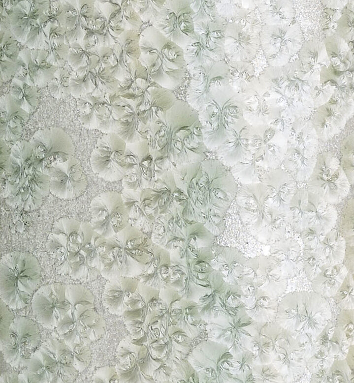 A close-up image of a delicate pattern featuring overlapping, soft white and light green floral motifs resembling hydrangea blooms. The flowers are intricately detailed, creating an elegant, textured surface.