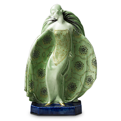 A ceramic figurine of a woman in an elegant, flowing green and gold dress stands on a blue base. Her arms are raised, and the dress features intricate patterns with an art nouveau style. Her head is tilted slightly, and her hair appears to be styled in an updo.