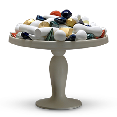 A beige pedestal cake stand holds an assortment of colorful, abstract ceramic shapes, including cylinders, cones, and spheres in various shades of gold, blue, white, and green. The arrangement creates a modern, artistic display.