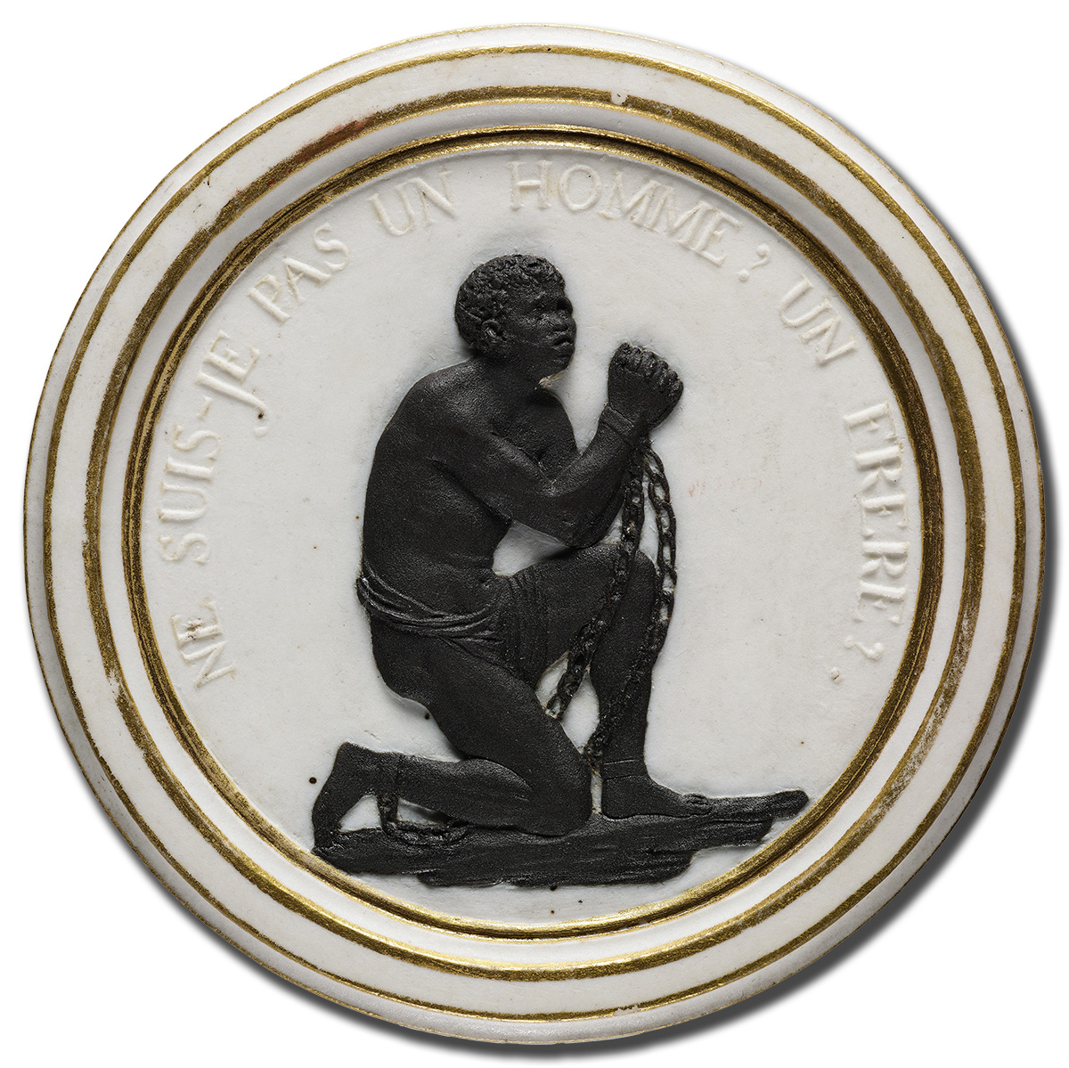 A circular medallion with a depiction of an enslaved person, kneeling with hands and feet shackled in chains. The text around the border reads, "NE SUIS-JE PAS UN HOMME? UN FRÈRE?" which translates to "Am I not a man and a brother?.