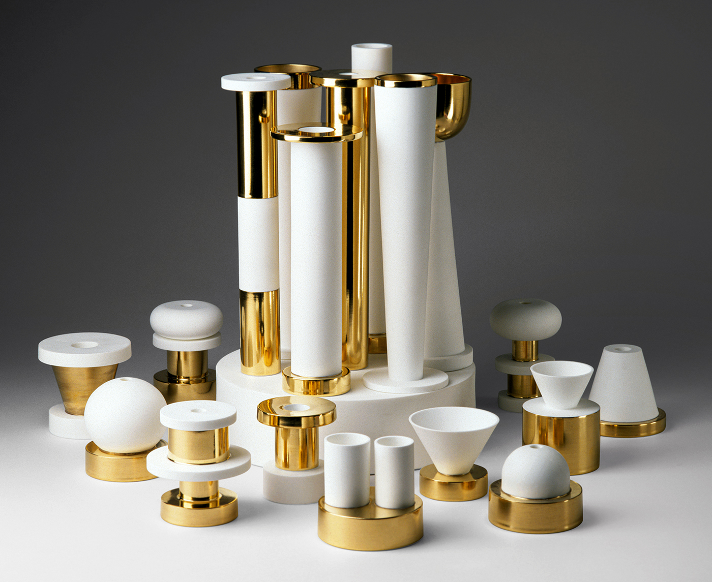 A collection of abstract, modern sculptures featuring cylindrical, spherical, and conical shapes in white with shiny gold accents. The assortment varies in size and is arranged on a neutral gradient background from light to dark.