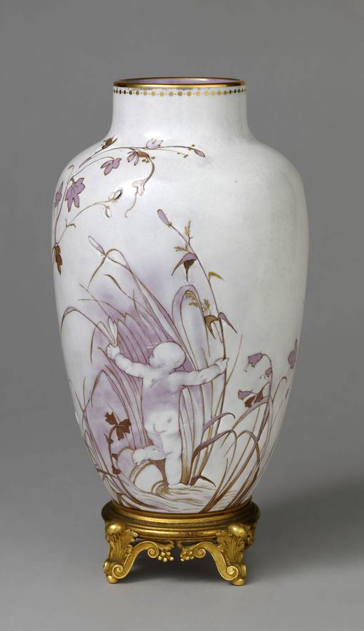 A tall, decorative porcelain vase adorned with a floral pattern featuring purple and gold hues. The vase sits on an ornate gold-colored stand with intricate designs. The background is a plain gray.