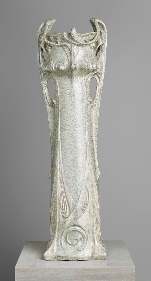 A tall ceramic vase with an intricate design features a greenish glaze. The vase has openwork handles and elaborate carvings with a nature-inspired, Art Nouveau style, including swirling motifs and organic forms. It stands on a pedestal against a plain background.