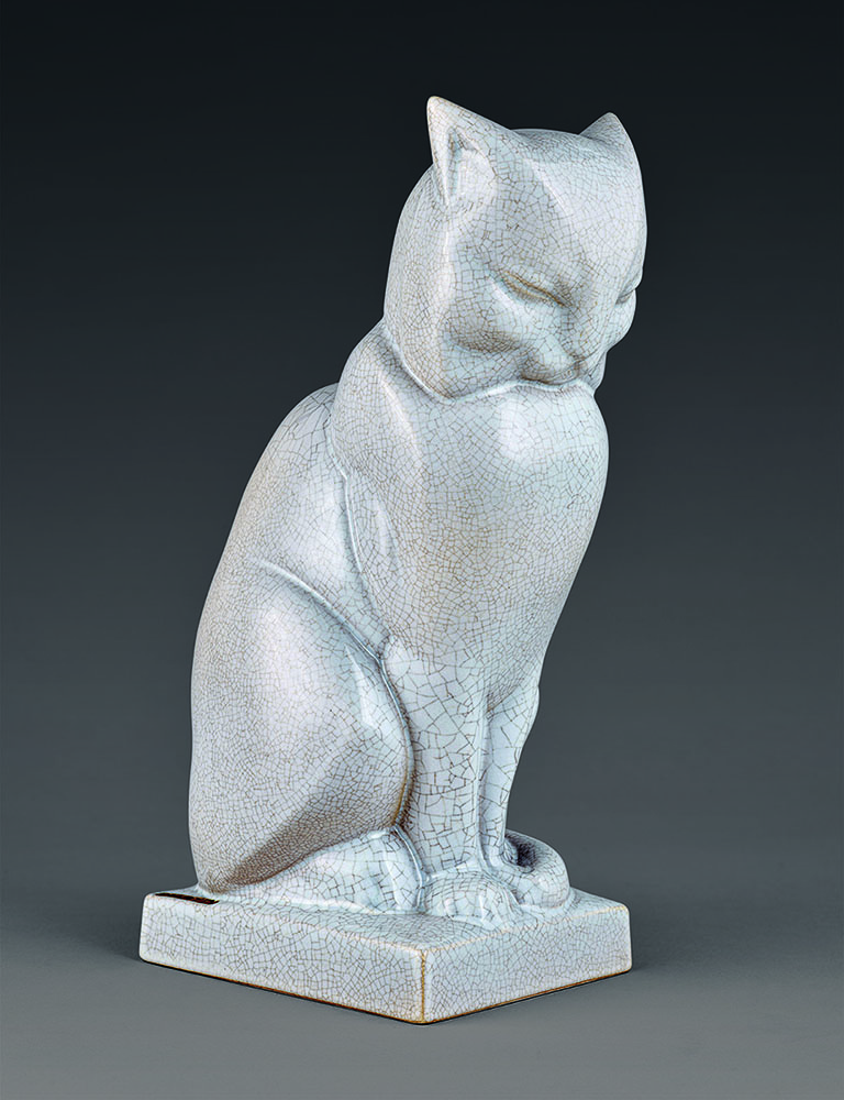 A ceramic sculpture of a cat sitting upright on a square base. The cat has a cracked, glazed surface with a light blue-gray color, and it appears to be looking downward, creating a serene and contemplative expression. The background is a plain dark color.