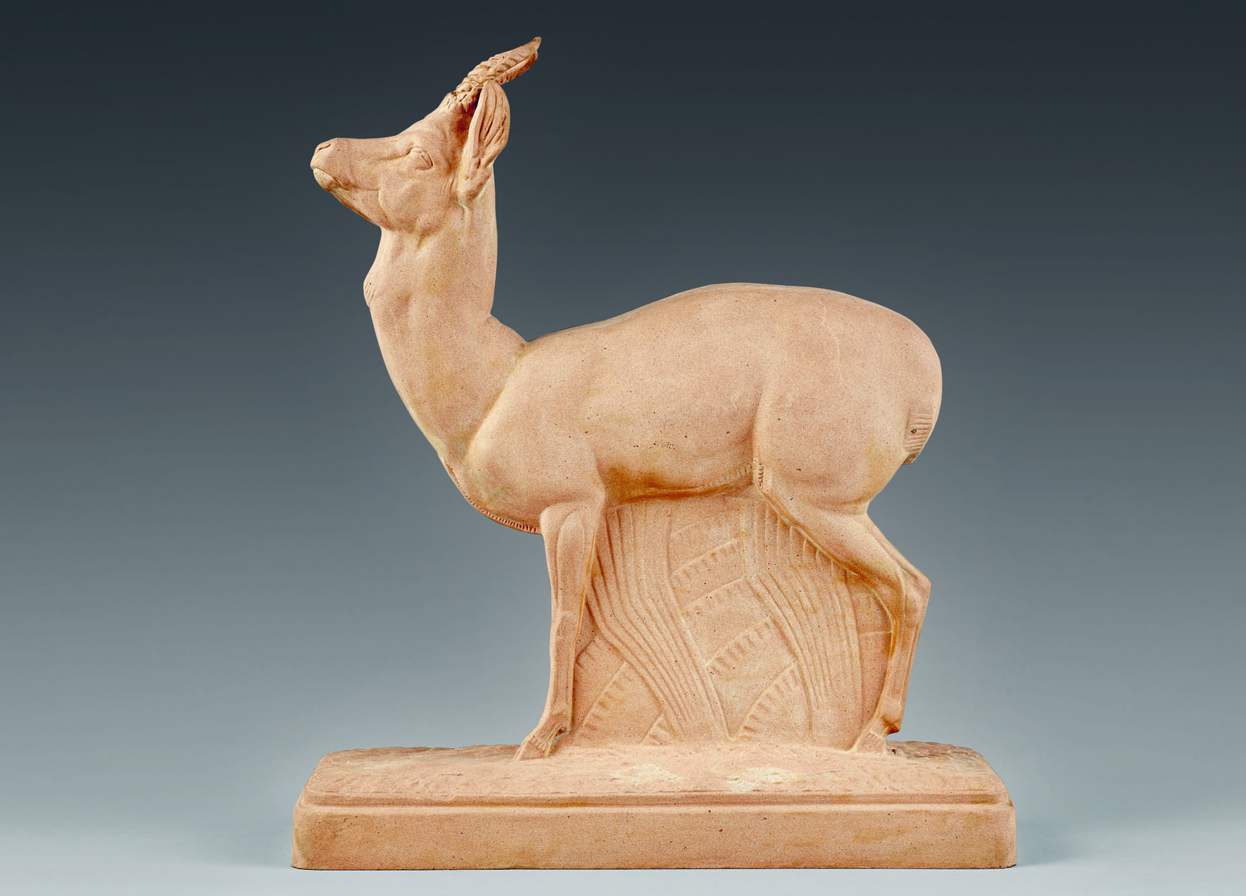 A beige sculpture of a standing antelope with its head tilted upward, mounted on a rectangular base. The animal's body is detailed with visible muscles and a textured background behind it, resembling foliage. The background is a solid gradient from dark blue to light blue.