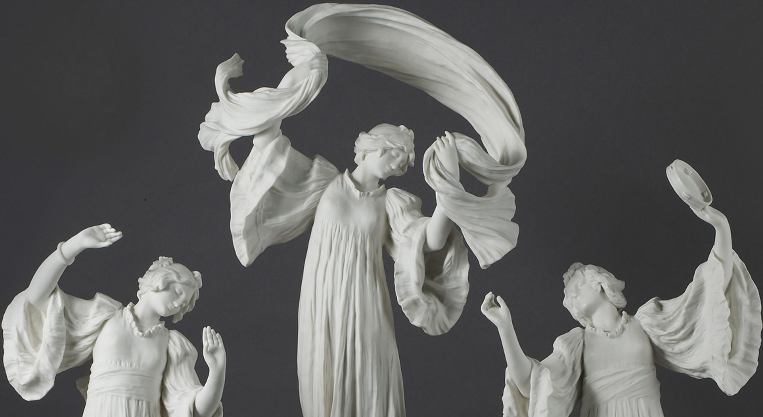 A detailed white marble sculpture of three women. The central figure stands with flowing garments and raised arms, holding a billowing cloth above her head. The two flanking figures mirror her gestures with their arms raised gracefully. The background is dark.