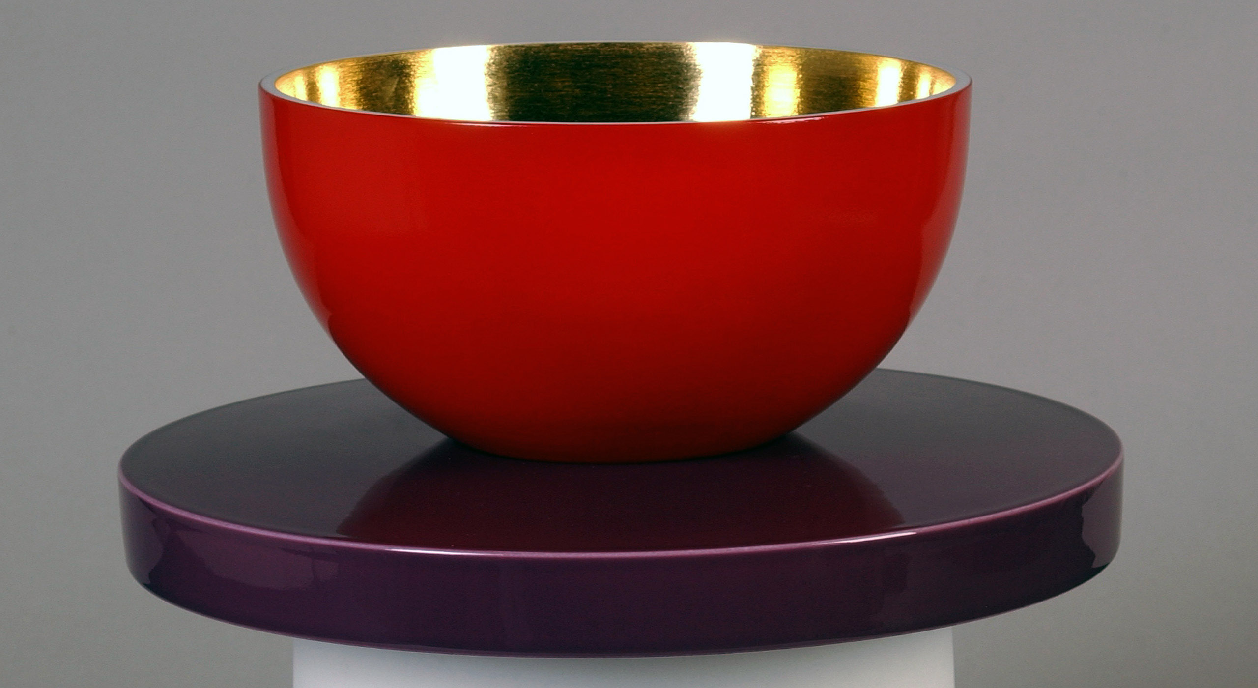 A polished red bowl with a gold interior sits on a glossy purple stand. The bowl and stand contrast in color, creating a striking visual effect, against a plain grey background.