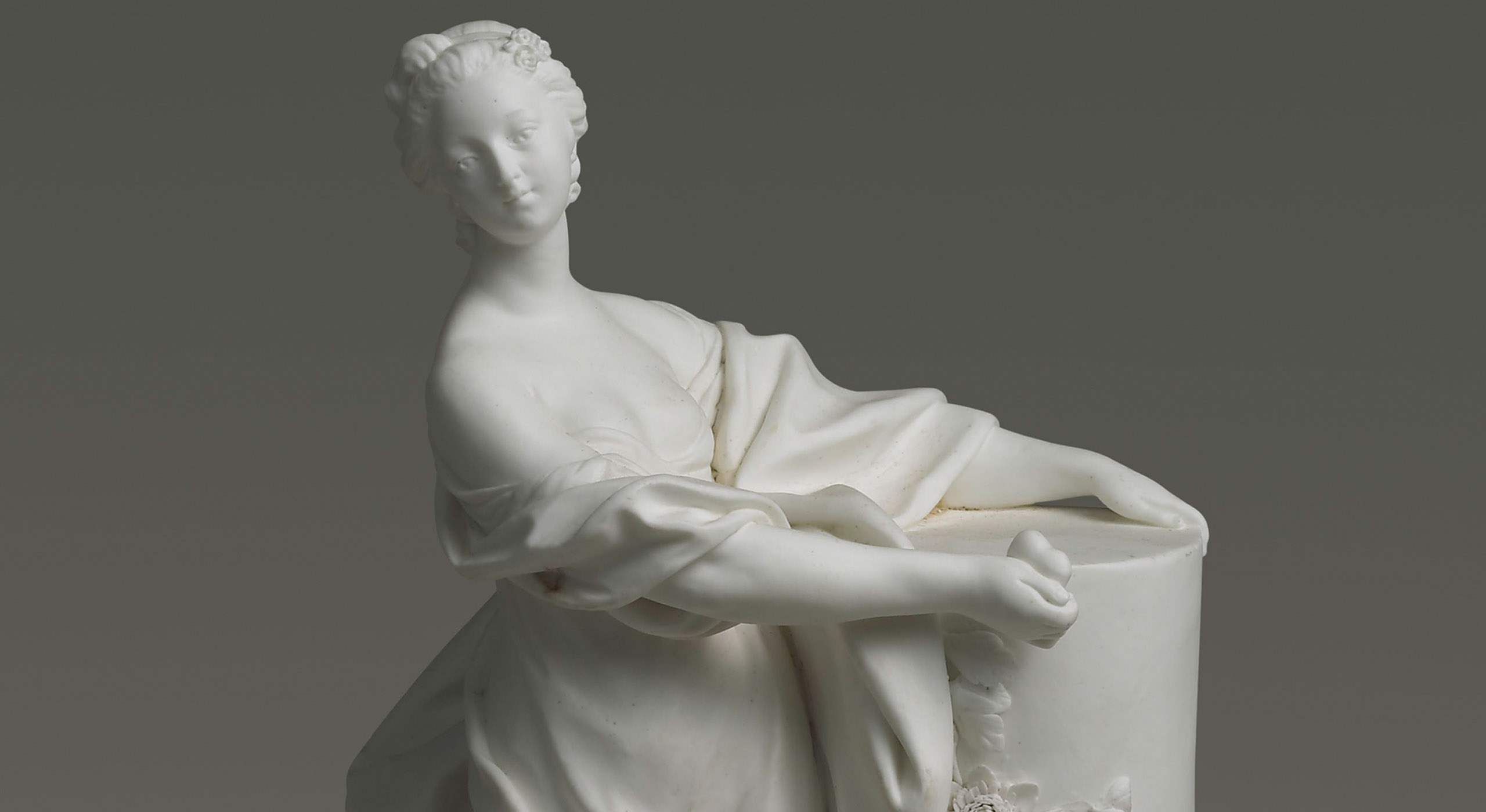 A detailed marble sculpture of a woman draped in a flowing fabric, gazing slightly to her left. Her left arm rests on a cylindrical pedestal adorned with carved flowers, while her right arm extends outward as if in motion. The background is a solid gray.