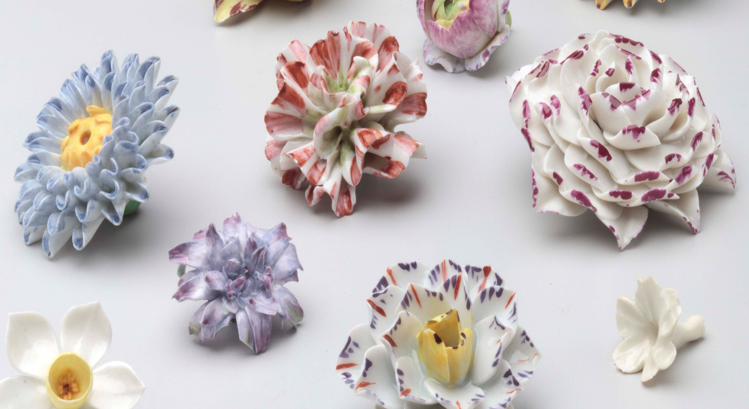 A collection of colorful ceramic flowers arranged on a plain white surface. The flowers vary in shapes and colors, including shades of purple, pink, yellow, and white, with intricate petal details. Each flower is distinct, lending a diverse display of floral art.