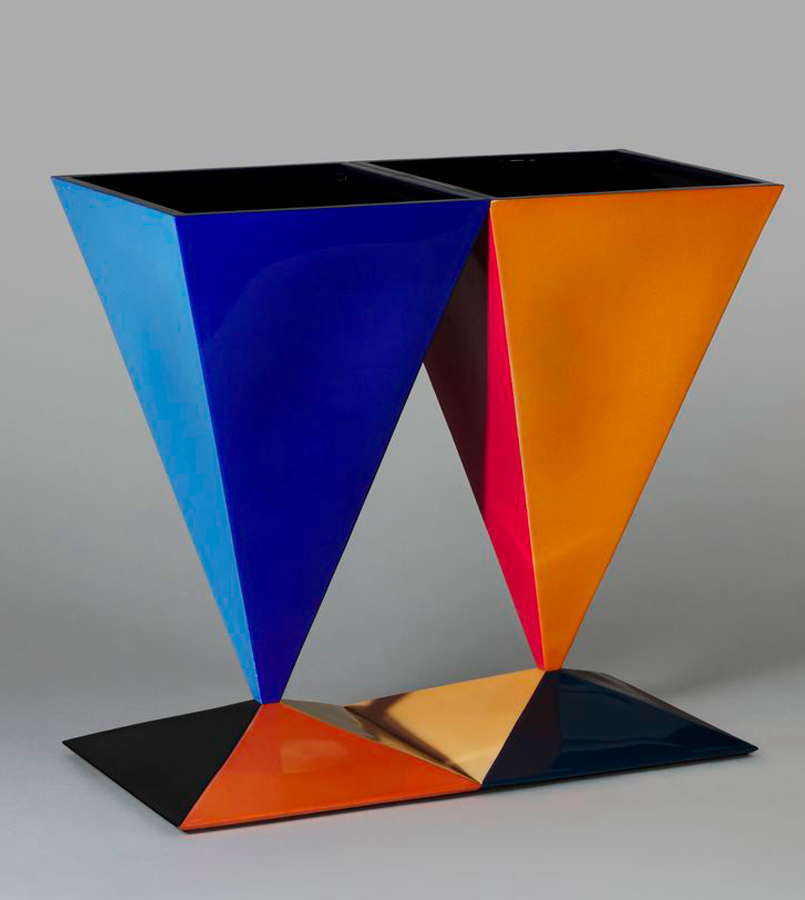 A colorful sculpture featuring two inverted pyramid shapes. One pyramid is blue and the other is a gradient of yellow, orange, and red. They are connected at their bases on a flat, rectangular platform and stand against a neutral grey background.