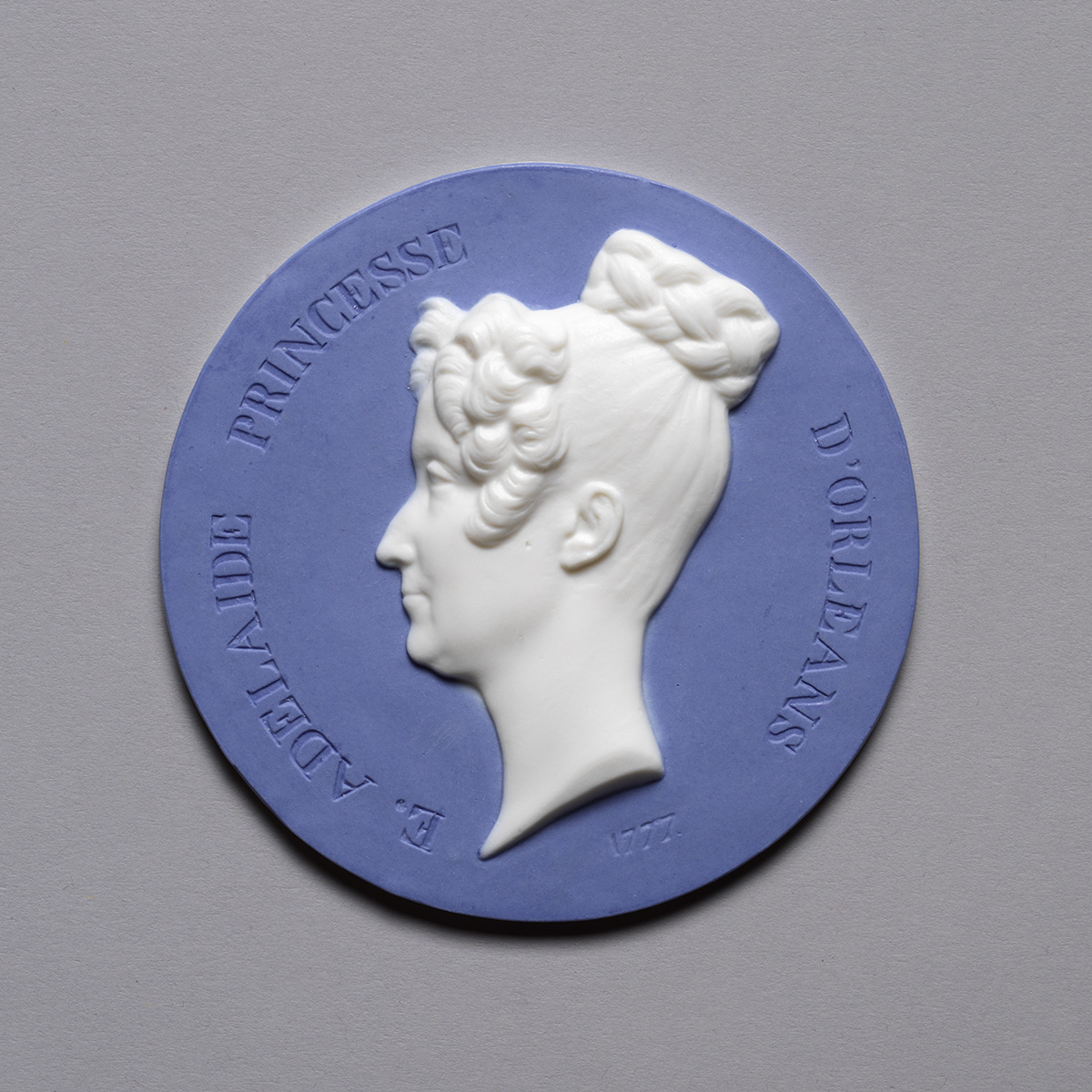 A circular ceramic medallion with a white profile of a woman's head against a blue background. The inscription reads "D'Adelaide Princesse D'Orleans" with the year "1777" at the bottom. The woman has a braided hairstyle.