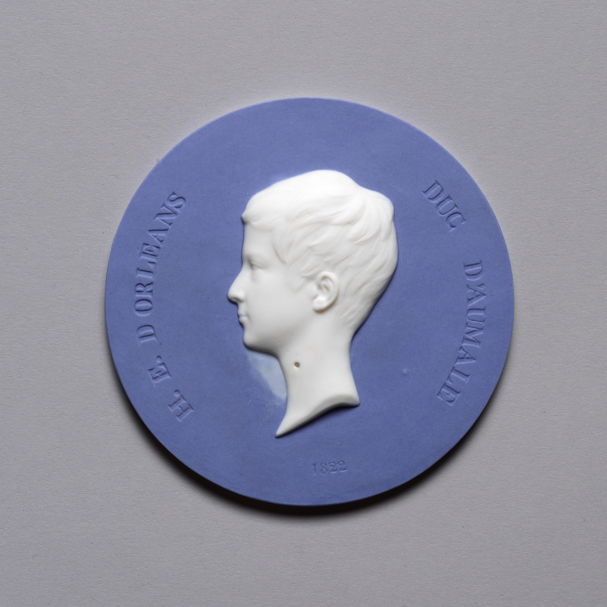 A round blue and white medallion featuring a profile of a young boy in white relief against a blue background. The surrounding text reads "H.L. D'Orleans Duc D'Aumale" with the year "1822" inscribed below.