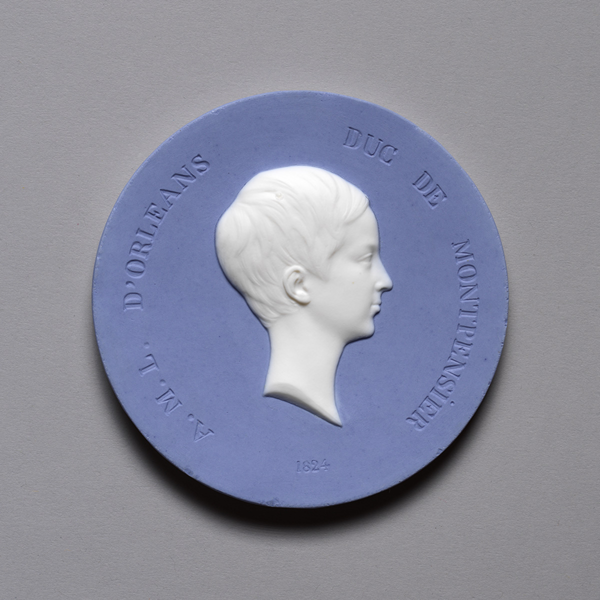 A circular medallion featuring a raised profile of a young person in white against a blue background. The inscription around the edge reads "FML. D'ORLEANS DUC DE MONTPENSIER 1824.