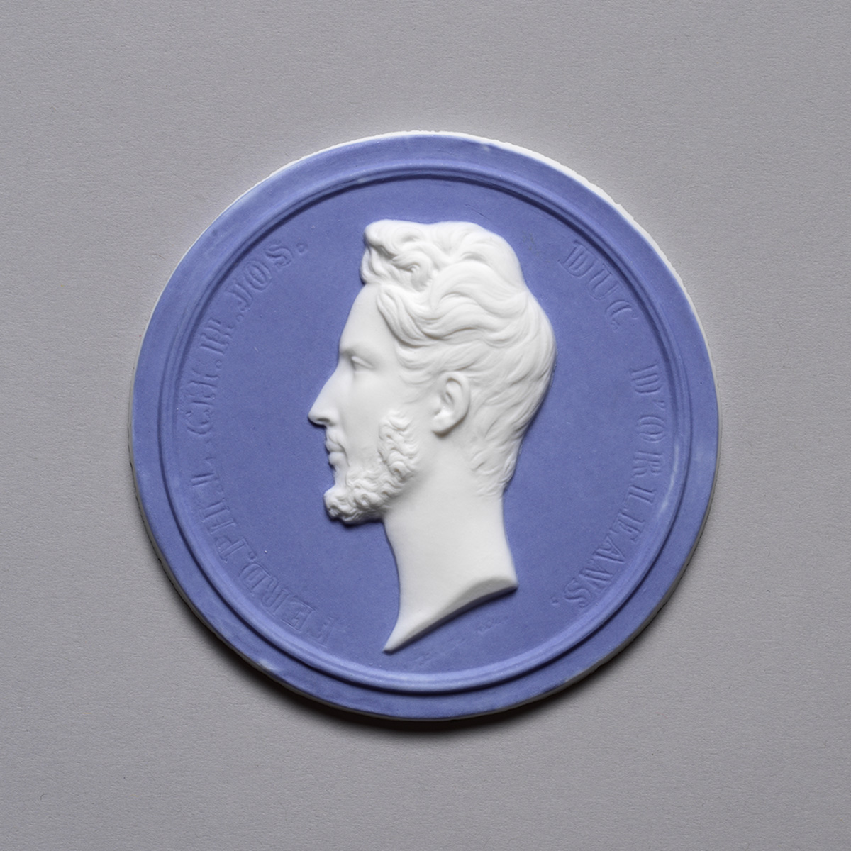 A blue and white circular medallion featuring a profile of a man with a beard and styled hair. The background is blue, and the figure is in white relief. Surrounding text is visible around the edge of the medallion.