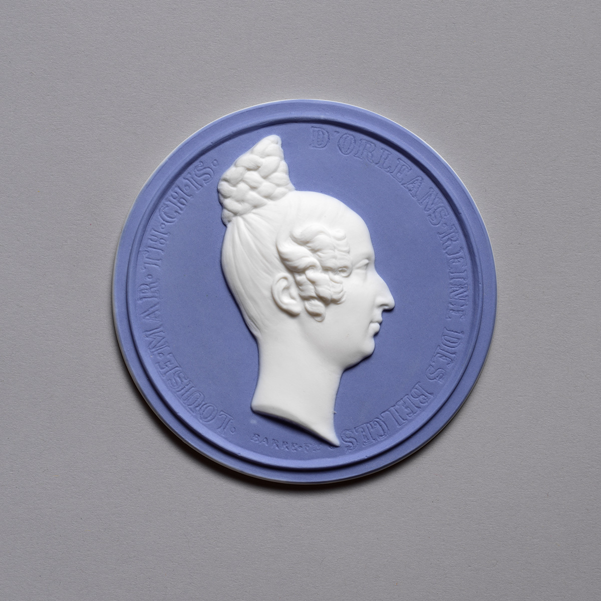 A round medallion with a profile of a woman with intricately styled hair in a bun. The medallion features raised white relief on a lavender background, with inscriptions around the edge.