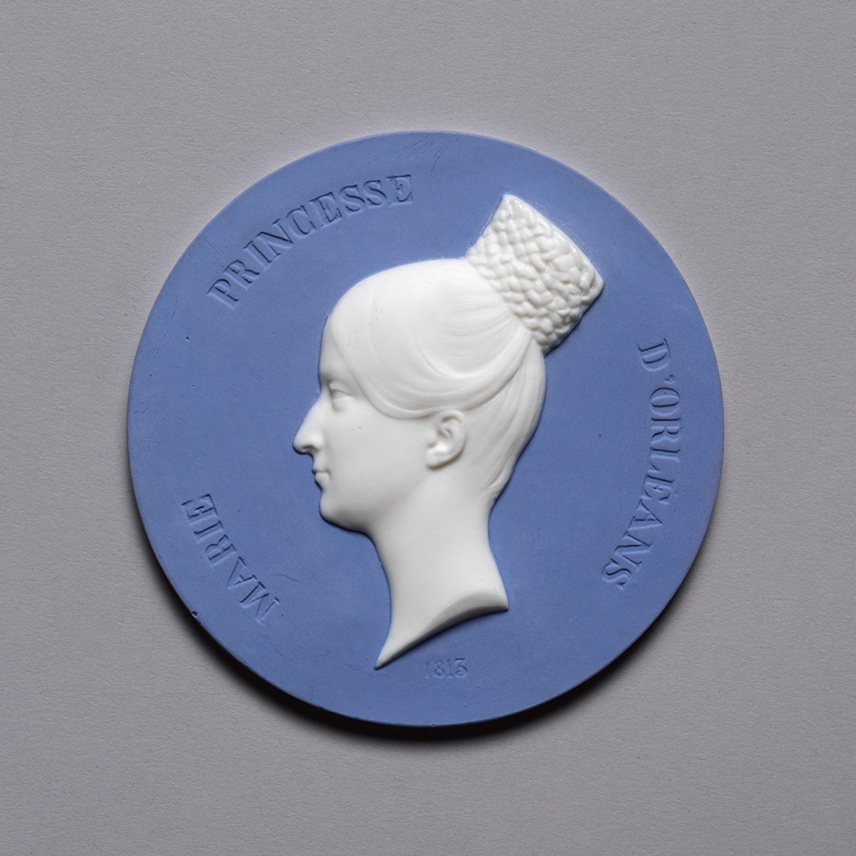 A circular blue and white medallion featuring a side profile of a woman with an elaborate hairstyle. Text around the edge reads "Marie Princesse d'Orleans" and the date "1815" is inscribed below the portrait.