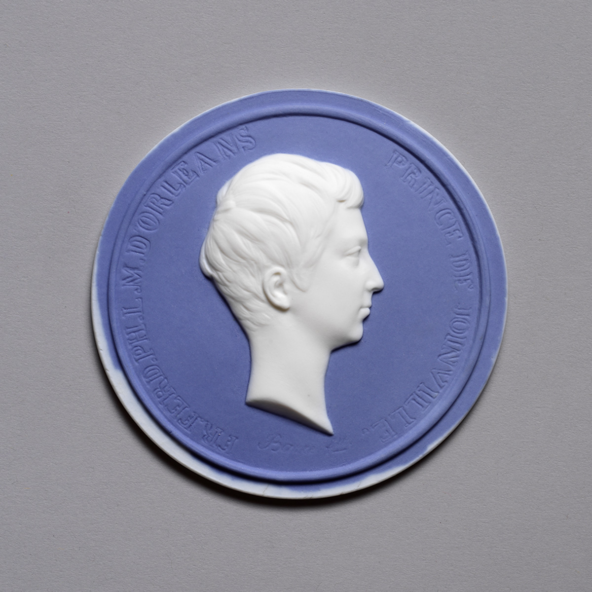 A round blue and white relief medallion featuring a side profile of a person's head. The surrounding text is embossed on the medallion. The background is a neutral gray.