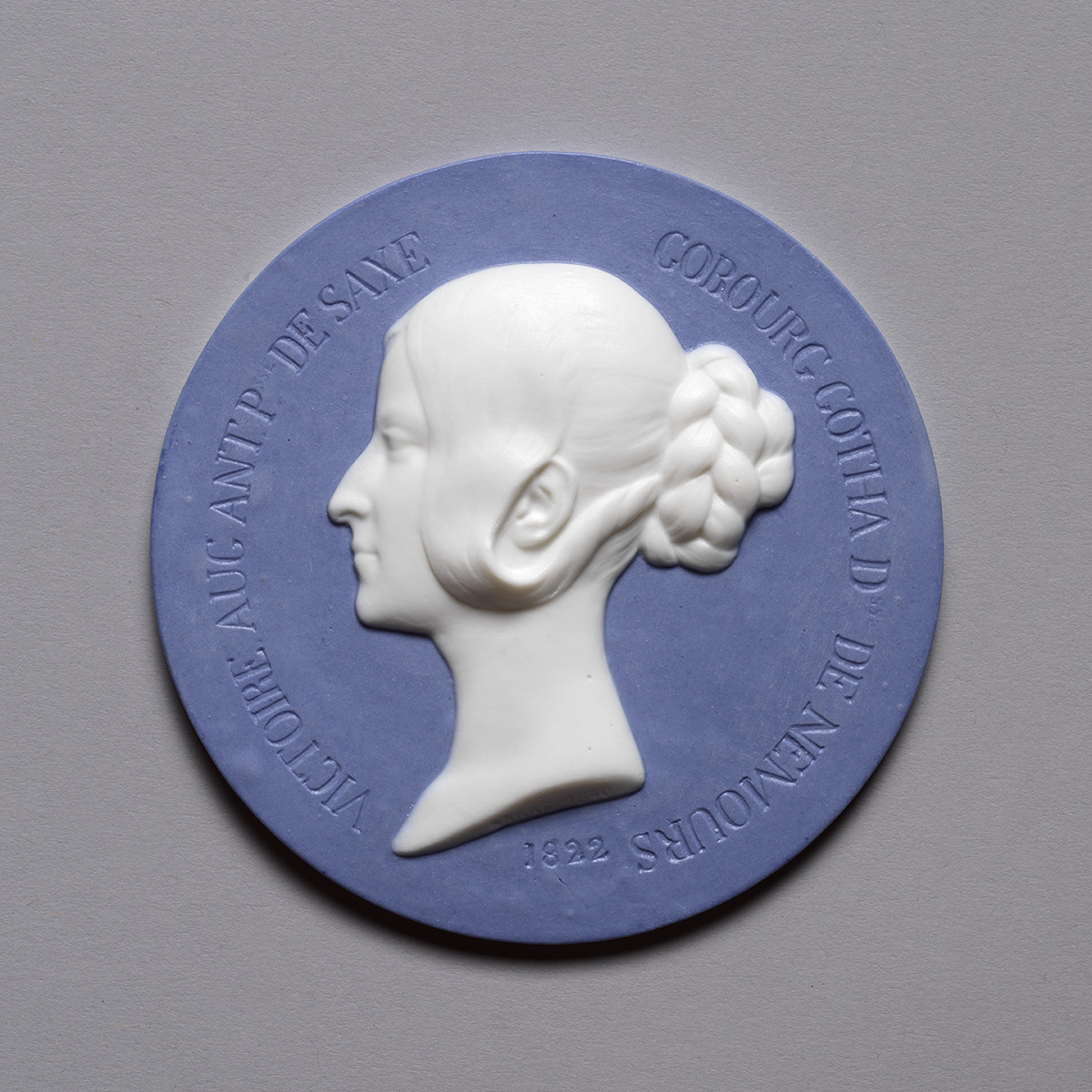 A circular blue medallion featuring a white relief of a woman's side profile with braided hair. The border has engraved text in French and the year 1822.