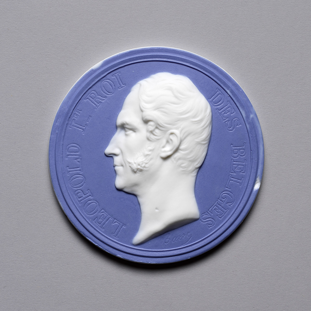 A blue and white circular medallion featuring a raised profile of a man facing left. Text around the edge reads "LEOPOLD 1ER ROI DES BELGES", with a small engraving at the bottom.