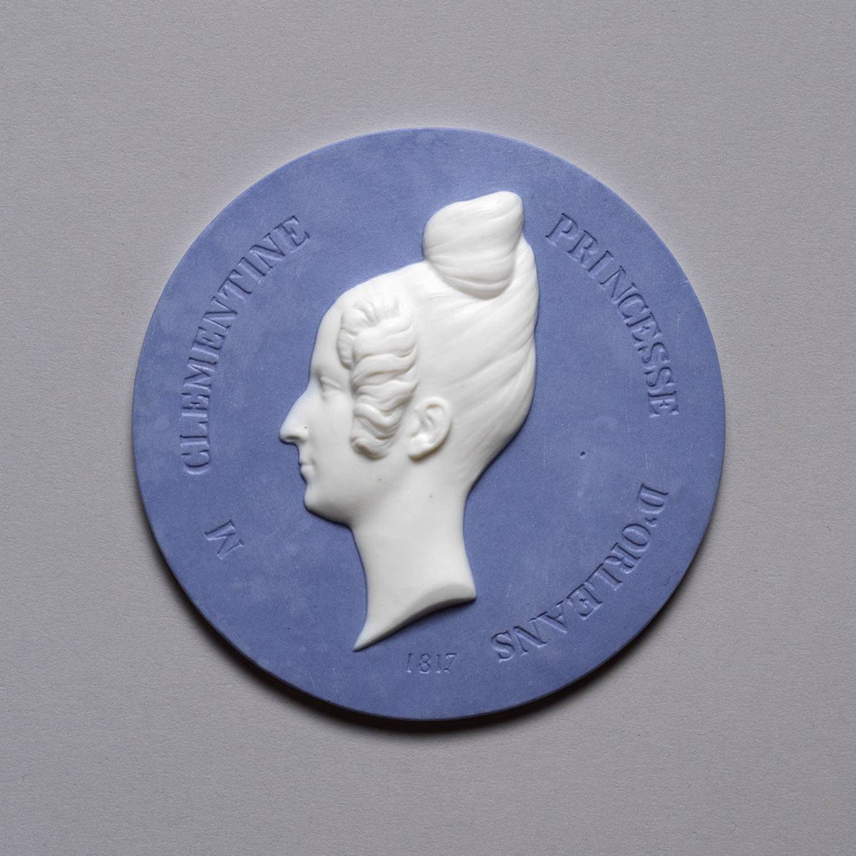 A blue medallion with a white relief profile of a woman wearing an intricate bun hairstyle, encircled by the inscription "CLÉMENTINE PRINCESSE D'ORLÉANS" and the year "1817.