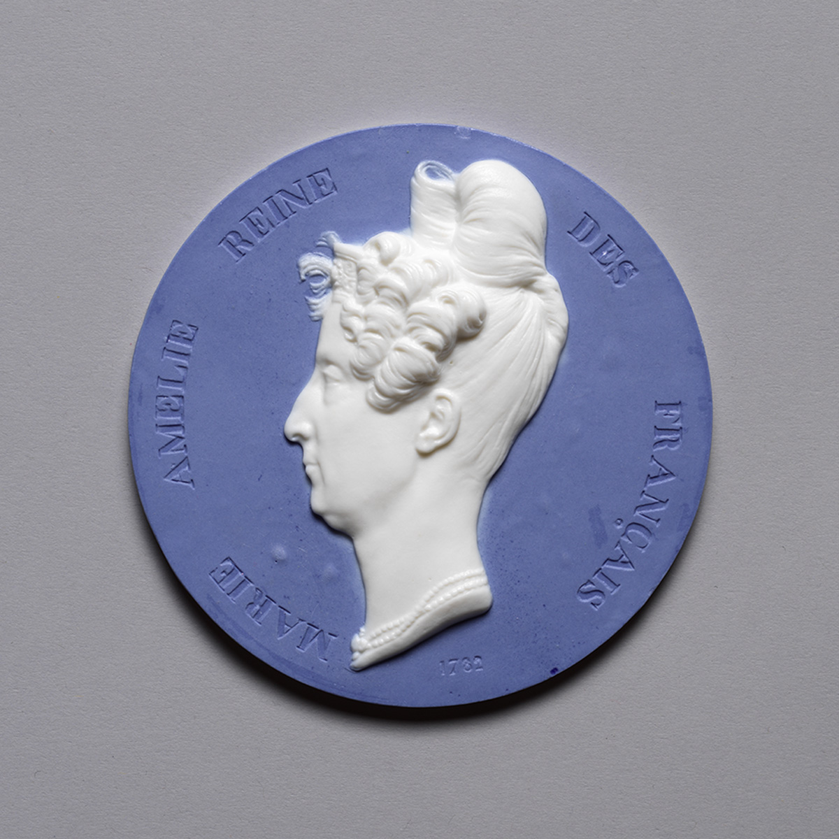 A round medal with a raised profile of a woman with an elaborate hairstyle. The inscription reads "Marie Amelie Reine des Français" against a blue background.