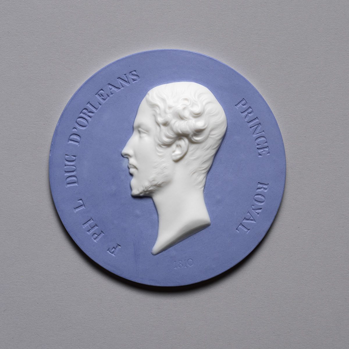 A blue and white medallion featuring a profile of a man with curly hair. The text around the edge reads, "F.EHL DUC D'ORLEANS PRINCE ROYAL" with the year 1830 engraved below the profile.